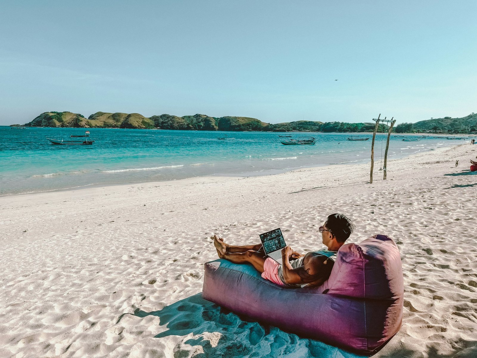 A man relaxing on a bean bag on a white-sand beach, working on his laptop