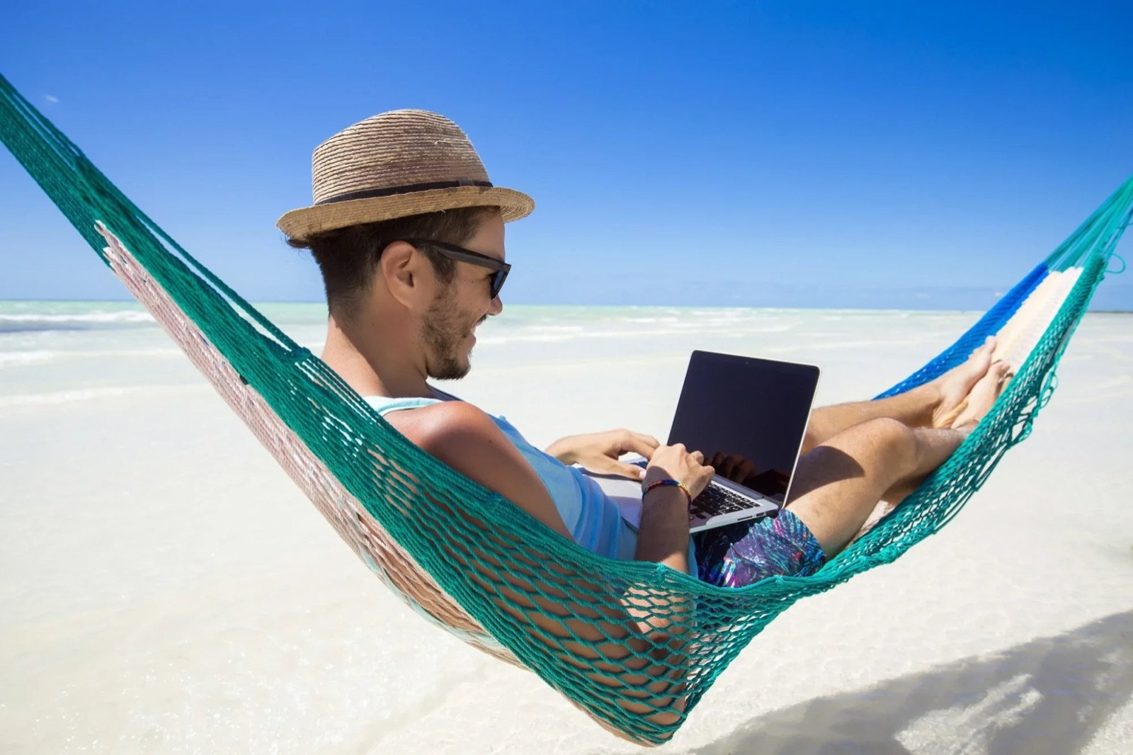 A smiling man in a straw hat working on a laptop in a hammock on a white-sand beach
